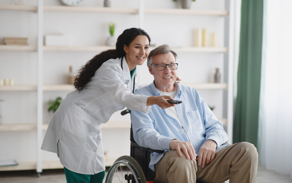 Young nurse with remote control switching channels on TV for senior man in wheelchair at retirement home